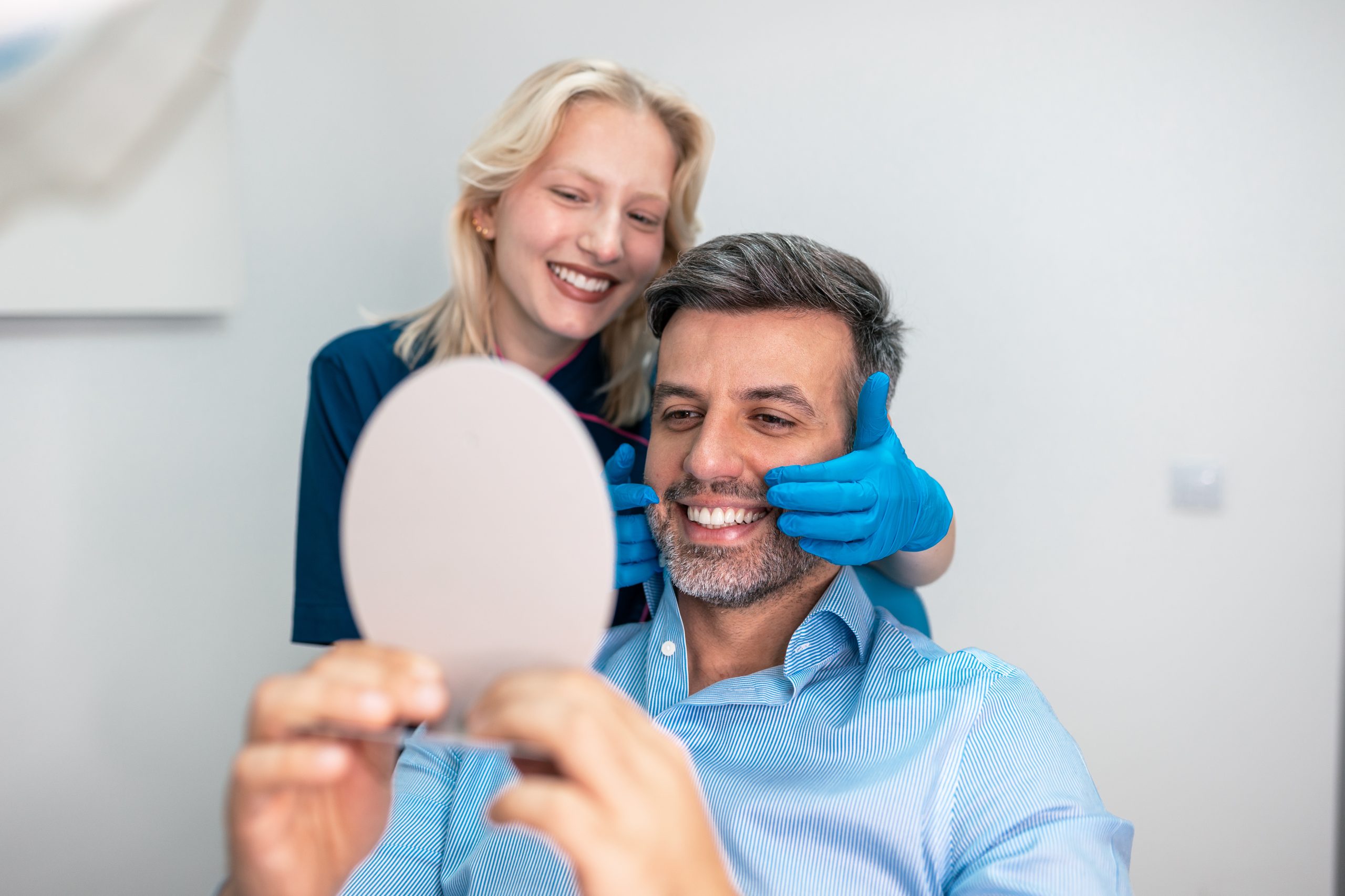 Happy man looking in mirror after dental treatment with female dentist, satisfied with his smile